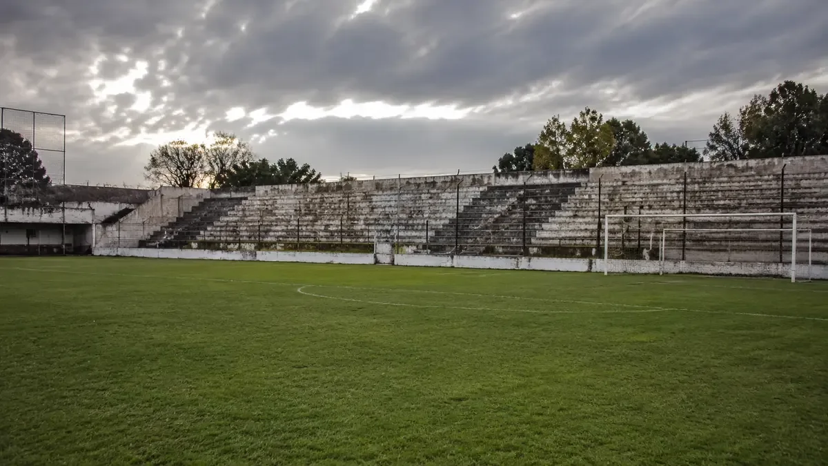 Cancha de fútbol de barrio con tribunas de cemento antiguas y desgastadas vacía